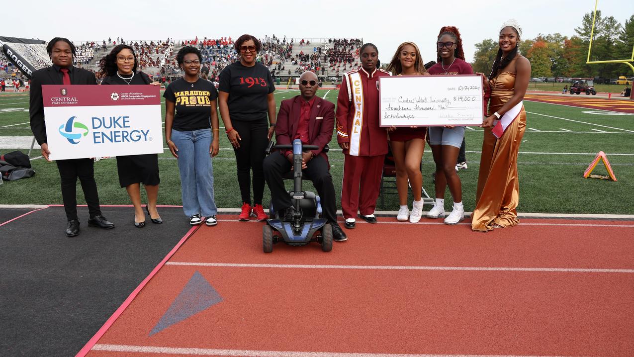 president kuti on the football field at homecoming while students hold large ceremonial checks from duke energy