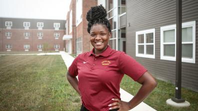 A young Black woman in maroon stands with hands on hips in front of residence halls at Central State University