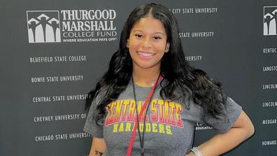 A young Black woman with long Black hair wearing a Central State University T-shirt in front of a backdrop for the Thurgood Marshall College Fund Yardi program