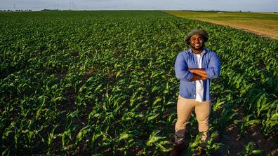 A young Black farmer stands with his arms crossed, smiling in a field of crops