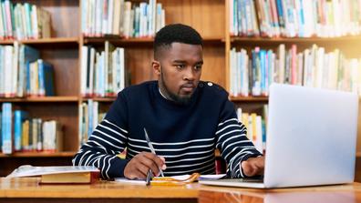 an African American student studies in a library using books and a laptop