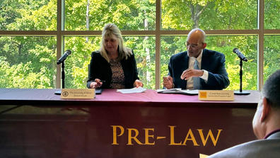 two people sit at a table with a tablecloth reading "Pre-Law" in front of windows with trees outside