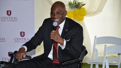 dr. morakinyo a.o. kuti wearing a black suit and maroon tie speaks into a microphone