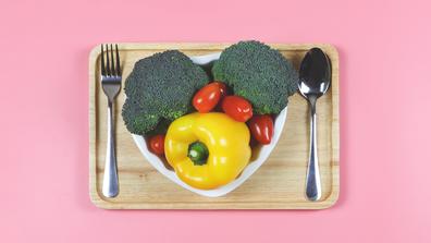 a heart-shaped plate filled with fresh vegetables with a spoon and fork on a pink background