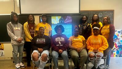 nine students and a professor in a central state university art classroom with a flyer for study abroad in paris on the screen behind them