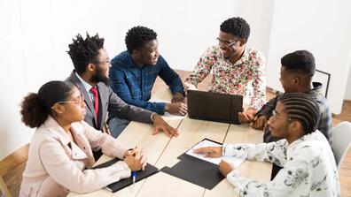 six students sit around a table with laptops and homework