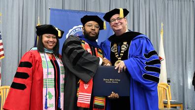 three people wearing regalia at a graduation ceremony with ramon moon in the center holding his diploma