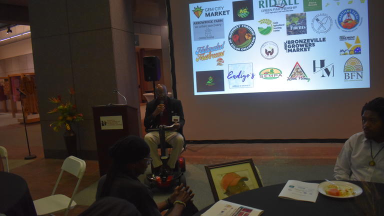 a person using a wheelchair speaks in front of a projector showing various logos