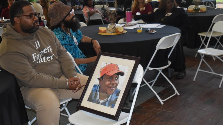 a person sits at a table beside an artist's rendition of a Black man in a red-orange hat