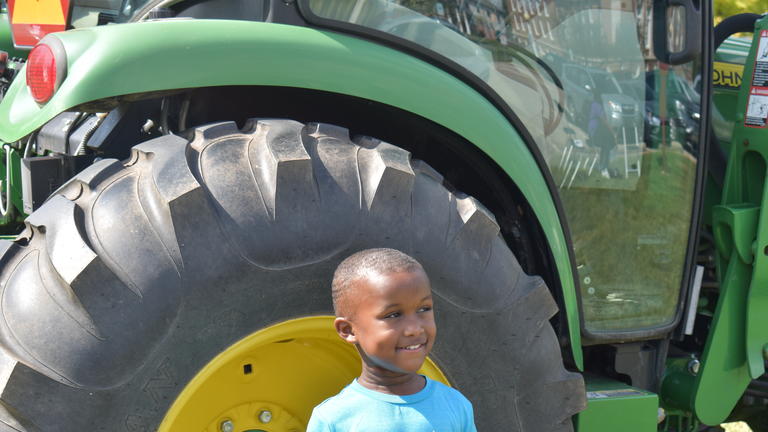 young child smiles to someone at his left while standing in front of a green tractor