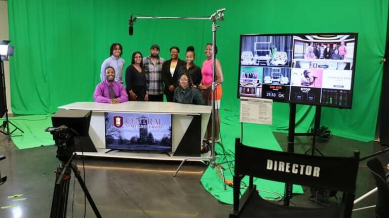 Students and Faculty Sitting at a desk in our inhouse film studio with greenscreen and director chair