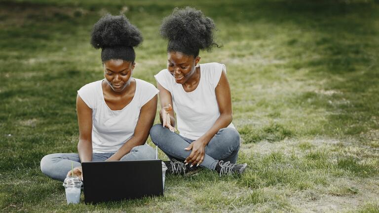 African American twin women sit in a grassy area with a laptop