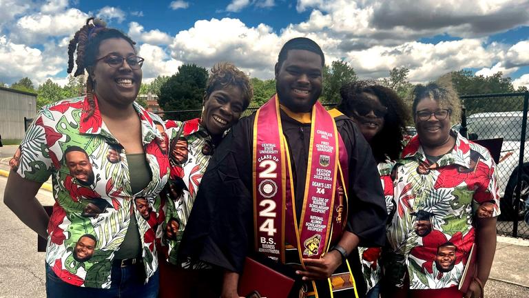 a graduate wearing regalia smiles with three people wearing shirts showing his photo