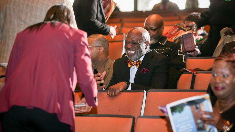 a man in a black suit smiles big while speaking with others at the convocation