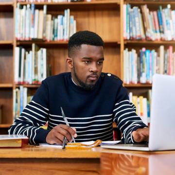 an African American student studies in a library using books and a laptop