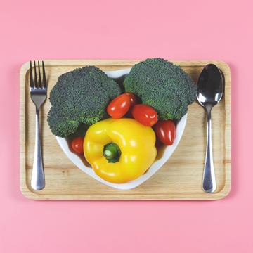 a heart-shaped plate filled with fresh vegetables with a spoon and fork on a pink background