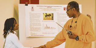 A student researcher shakes hands with an alumni attendee of Scholarly Research Activities Day at Central State University. The title of the research project shown on a posterboard in the background is, "Viral Prevalence of Small Carpenter Bees Among Different Farm Landscapes"