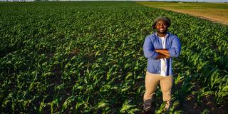 A young Black farmer stands with his arms crossed, smiling in a field of crops