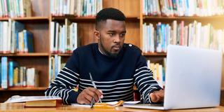 an African American student studies in a library using books and a laptop