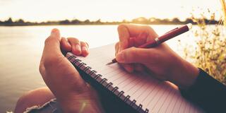 a person writes with a pen in a notebook beside a body of water