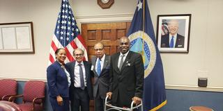 four people stand in front of a U.S. flag in a conference room