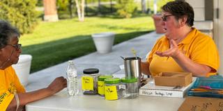 two people in gold shirts talk at a table with jars of seeds