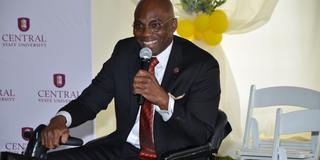 dr. morakinyo a.o. kuti wearing a black suit and maroon tie speaks into a microphone