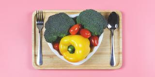 a heart-shaped plate filled with fresh vegetables with a spoon and fork on a pink background