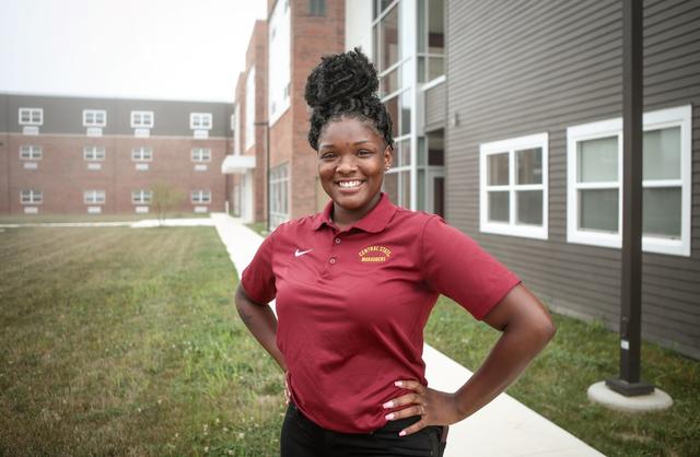 A young Black woman in maroon stands with hands on hips in front of residence halls at Central State University