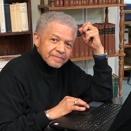 A Black man with gray hair wears all grey while smiling at the camera. He is using radio equipment.