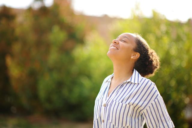 A young African American women looks up with her eyes closed, taking in the sunshine with greenery behind her