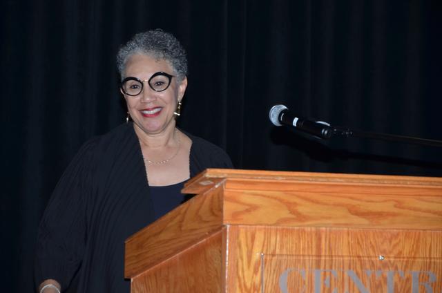 a black woman in black with classes smiles at a podium with a microphone