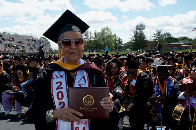 an older woman wearing sunglasses and graduation regalia holds her central state university diploma during commencement