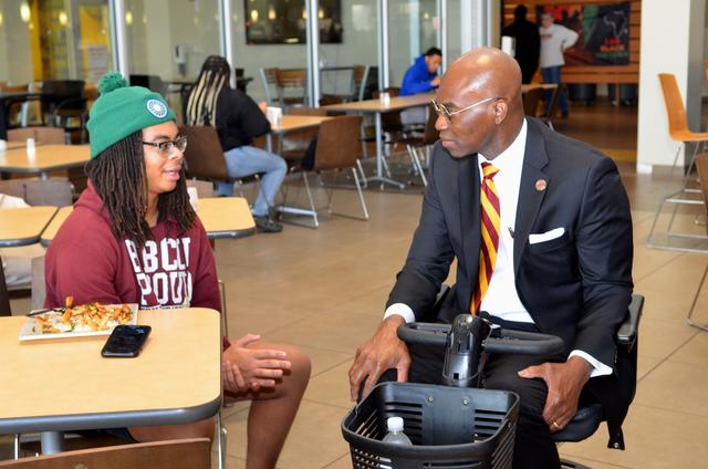 dr. morakinyo kuti, who uses a power chair, talks with a student wearing an hbcu proud hoodie and a green beanie at a table in the marauder cafe