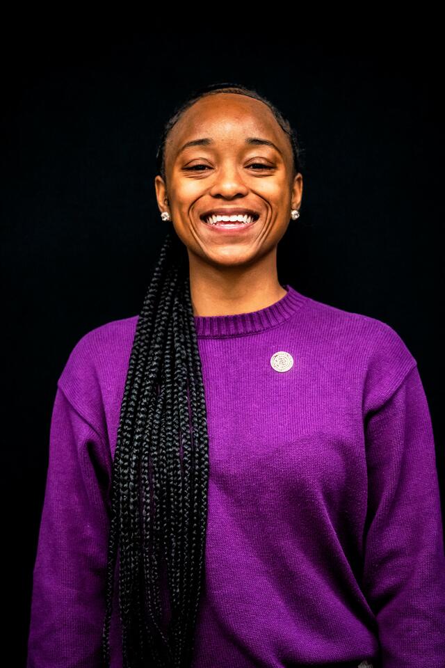 headshot of kamry hill, a young Black woman with locs wearing a purple sweater