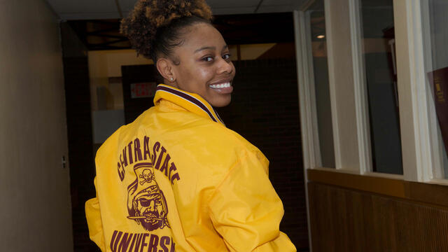 a central state university marauder looks back over their right shoulder, smiling at the camera. they are wearing a central state jacket in maroon and gold